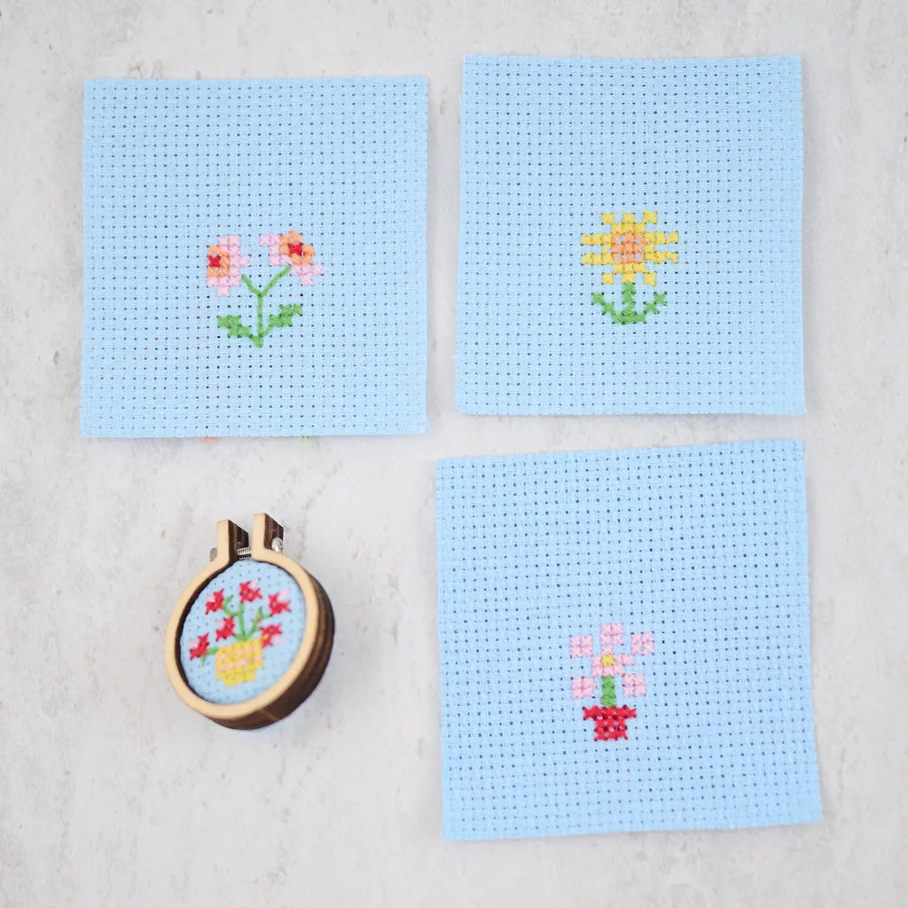 Image of all 4 possible cross stitch patterns in the mini matchbox cross stitch kit. All are on blue Aida cloth. The first is two pink flowers with red centers and green leaves, the second is a yellow sunflower, the third is in the mini hoop: six red roses in a tellow pot, the last is a pink daisy in a red pot. 