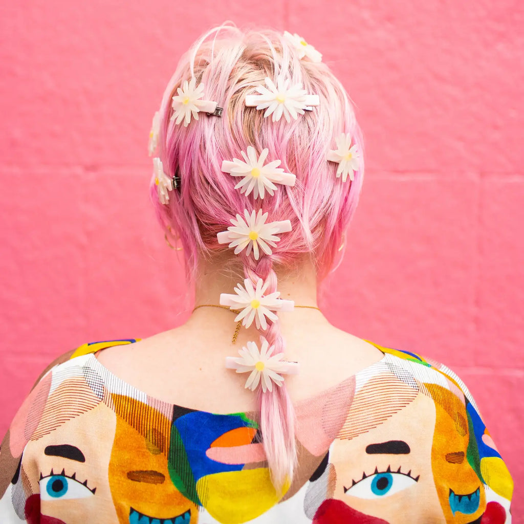 image of light skinned person with pink braided hair wearing 10 daisy hair clips all over their head with a colorful shirt with eyes on it against a pink brick background