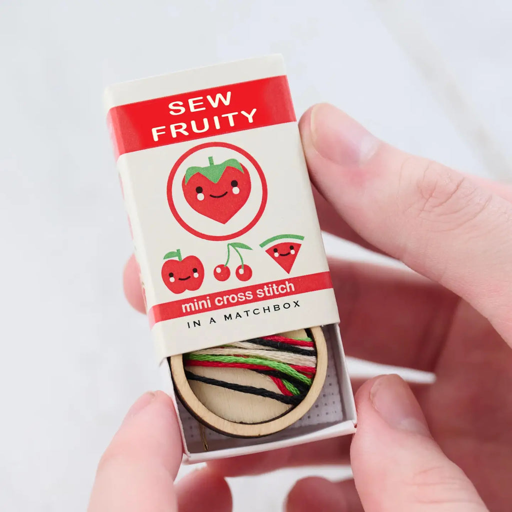 image show a matchbox containing a cross stitch kit with four different options of fruit to stitch: a strawberry, an apple, two cherrie, or a slice of watermelon - all using red and green. 